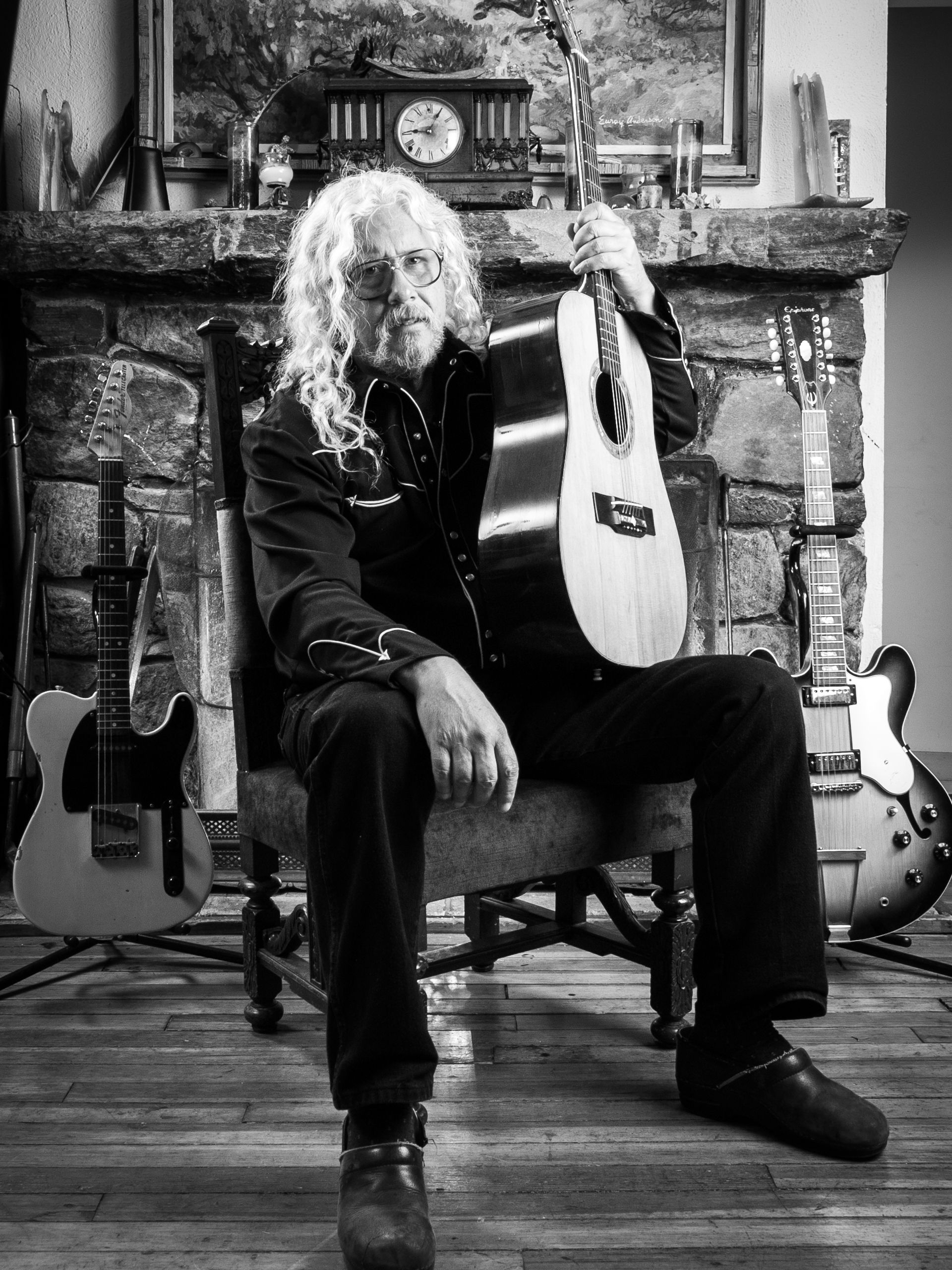 B&W portrait of Arlo Guthrie with his guitar sitting by his home fireplace