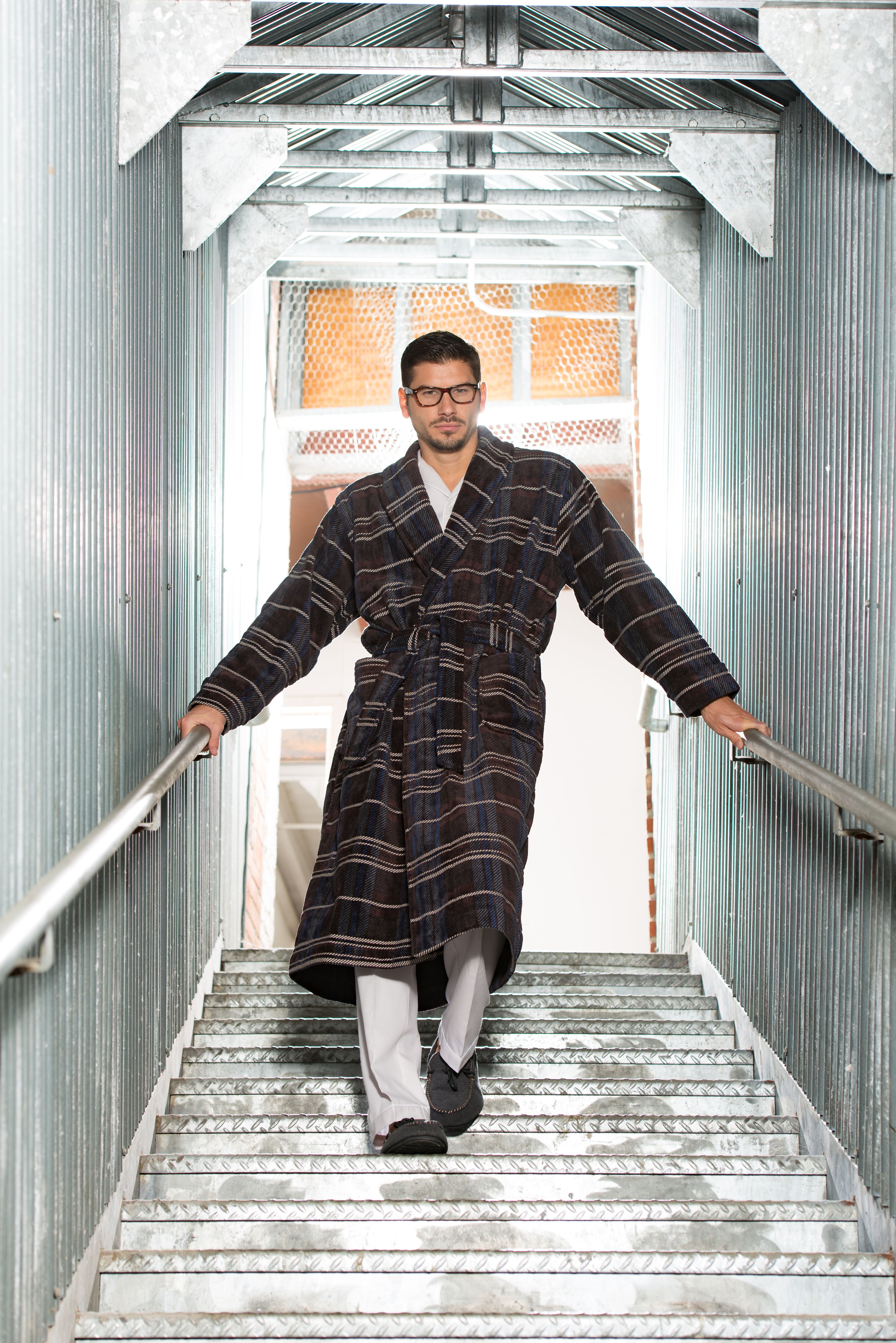 Male model in glasses and plaid bathrobe stands in silver stairwell at Mass MoCA in North Adams, Massachusetts