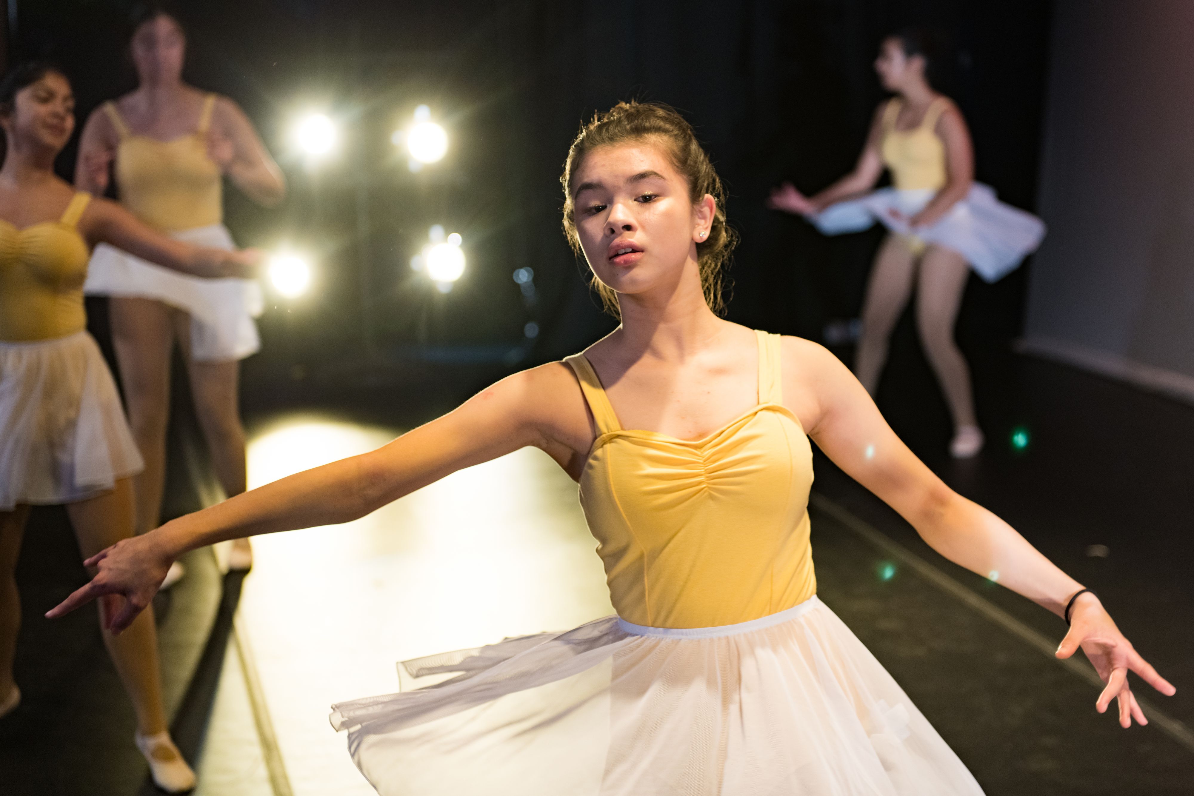 Young Ballerina on stage wearing yellow and white with three others and lights in background
