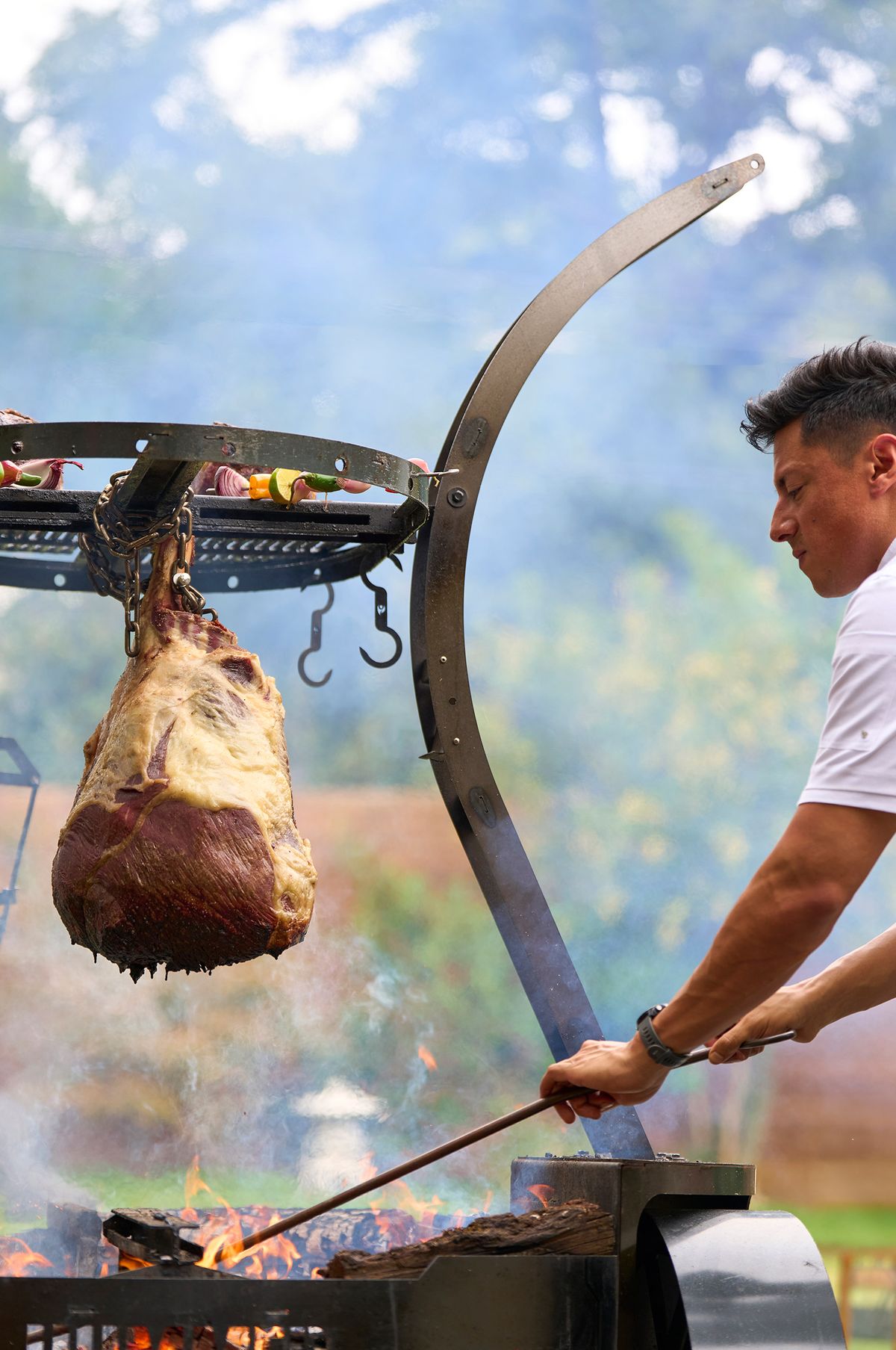 Chef tending the coals beneath a hanging Rosewood Wagyu cut, smoke rising across an outdoor live fire setup