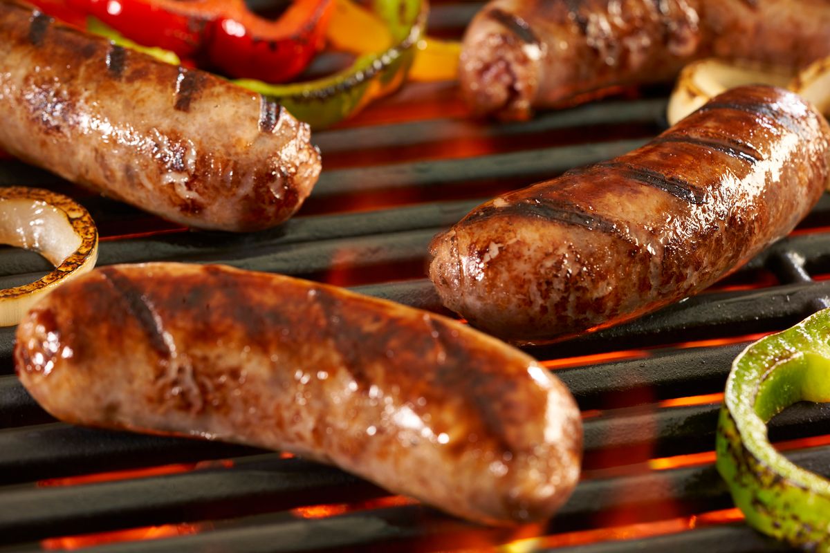 Bratwurst sausages searing on a hot grill grate with charred onion and bell pepper rings over glowing coals