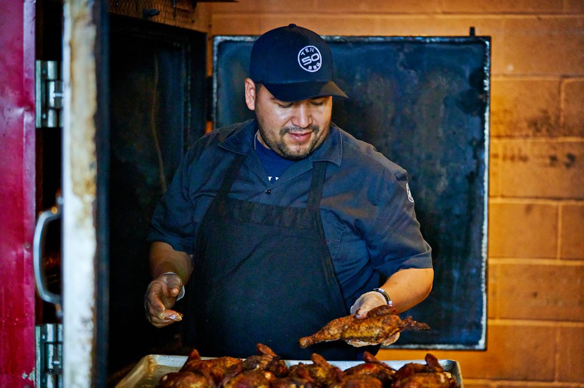 Ten50 BBQ pitmaster lifting a smoked half chicken from a full tray at the open smoker door