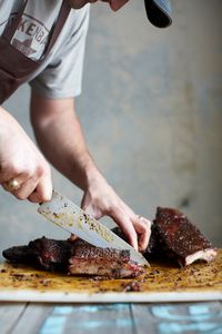Slicing a rack of Pecan Lodge pork ribs with a chef's knife on a cutting board, dark bark against a pale wall
