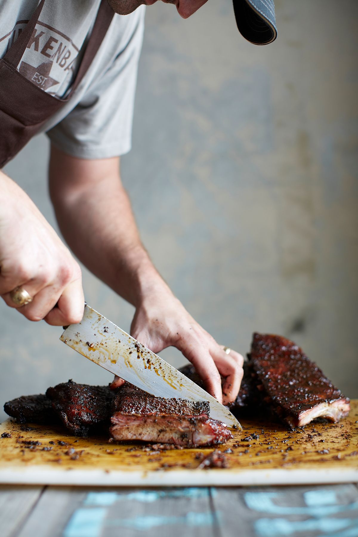Slicing a rack of Pecan Lodge pork ribs with a chef's knife on a cutting board, dark bark against a pale wall