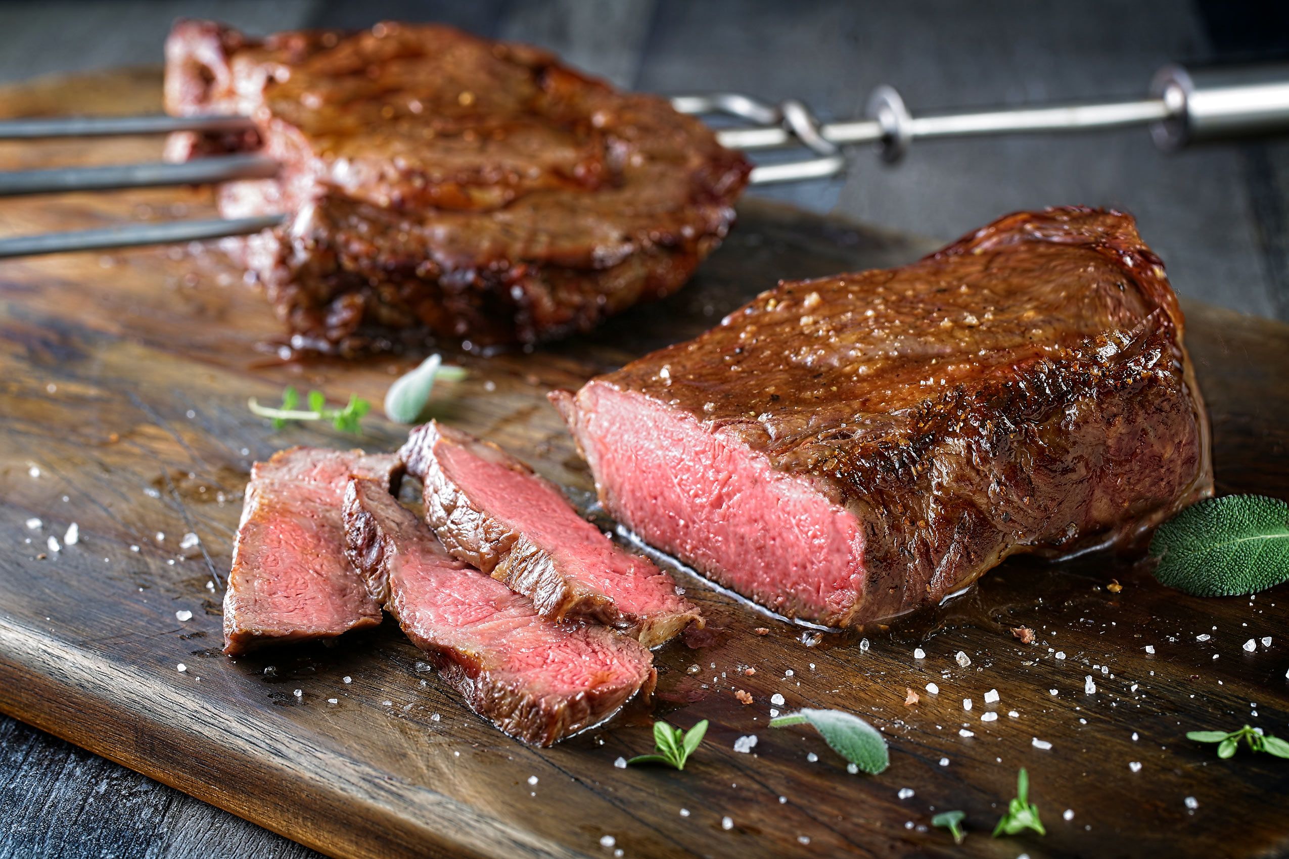Fogo de Chão steak sliced on a dark wood board with flaked salt, sage, and churrasco skewers behind