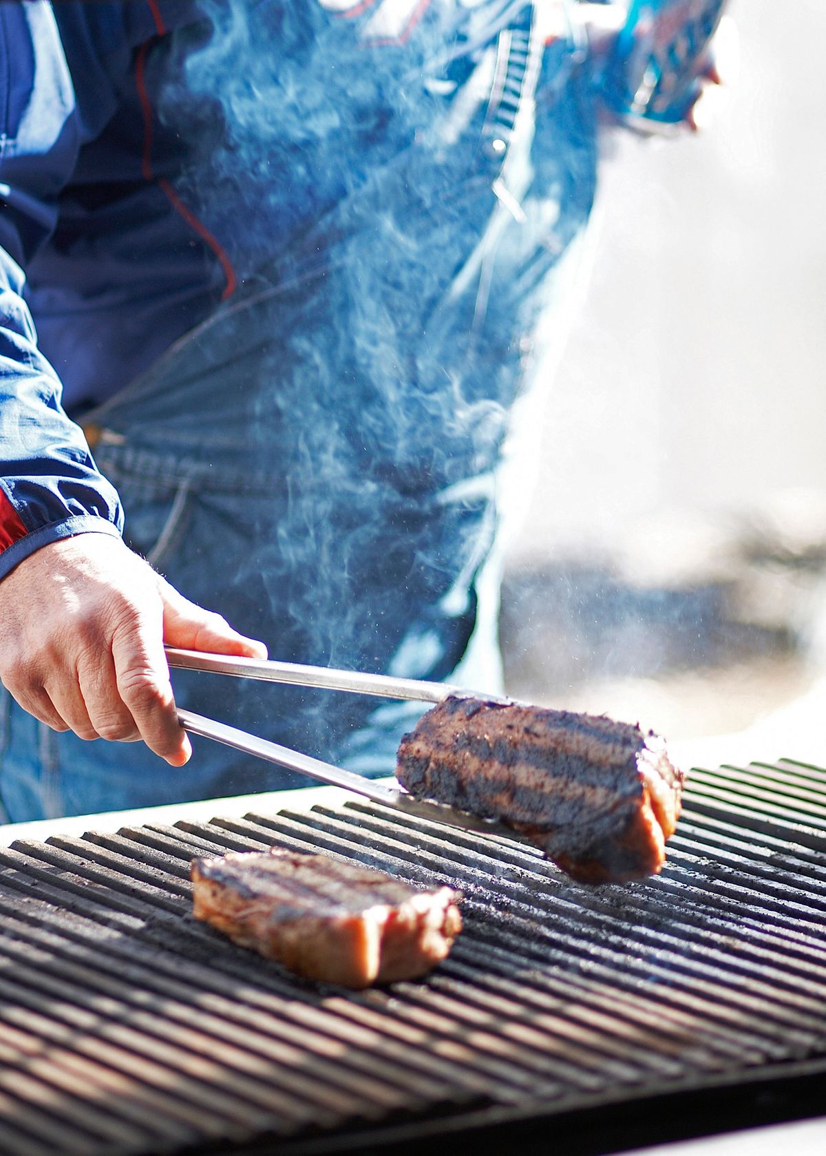 Turning grill-marked steaks with tongs over an open grate in bright sunlight at North Main BBQ