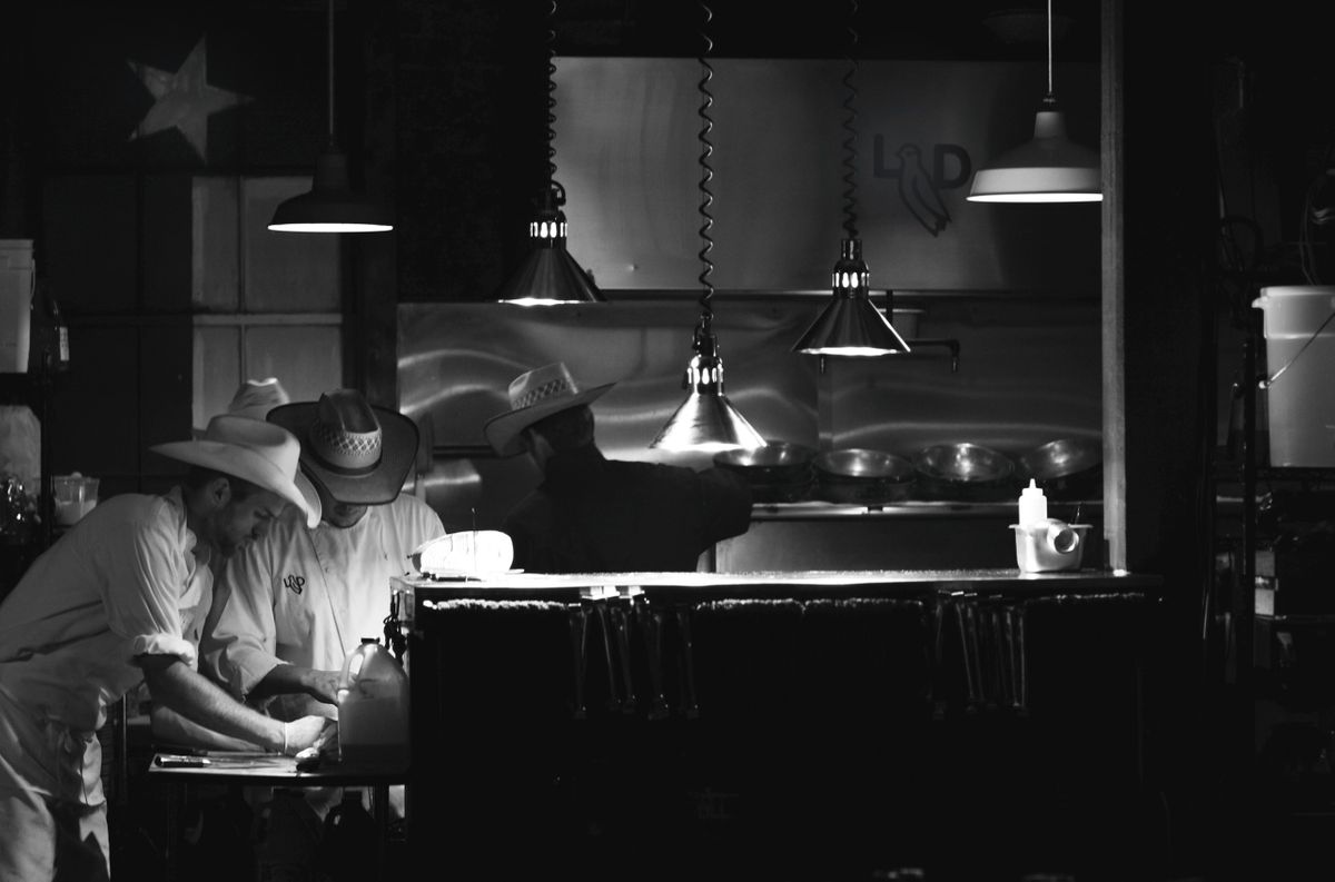 Wide black and white view of the Lonesome Dove kitchen line, cooks in cowboy hats beneath hanging heat lamps and the house logo