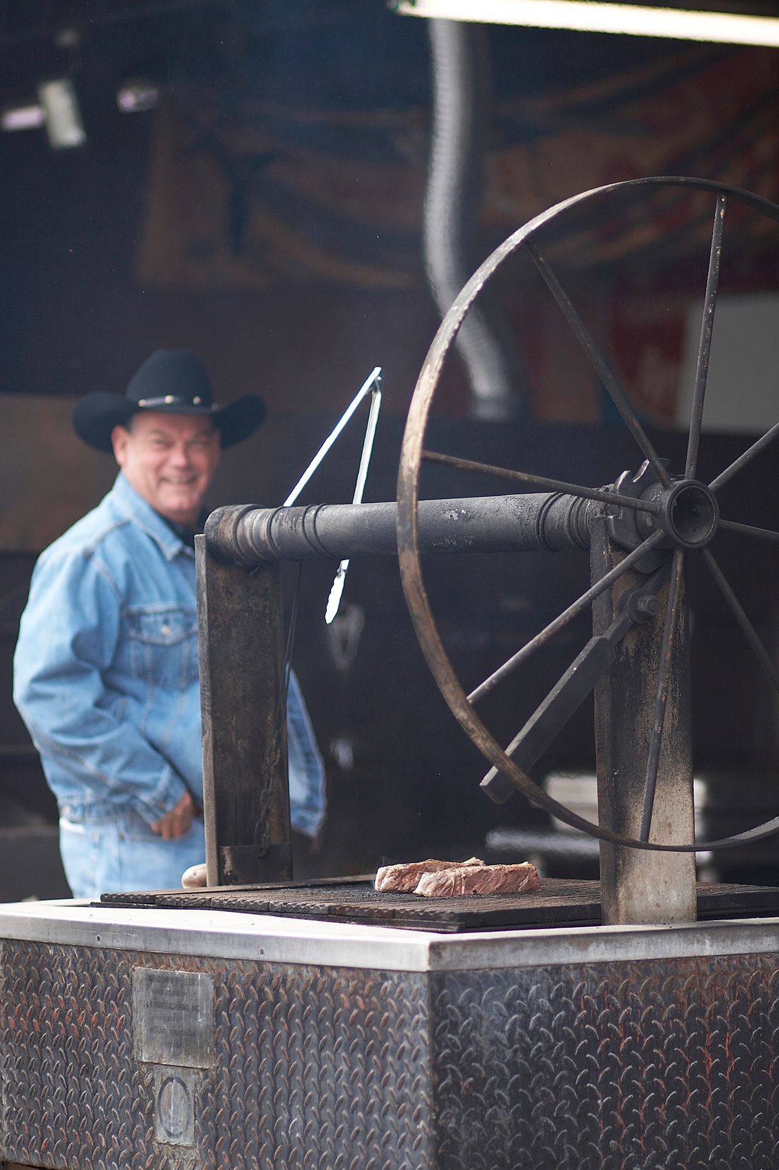 Pitmaster in a cowboy hat beside a crank-wheel grill with a single steak on the grate at North Main BBQ in Euless, Texas
