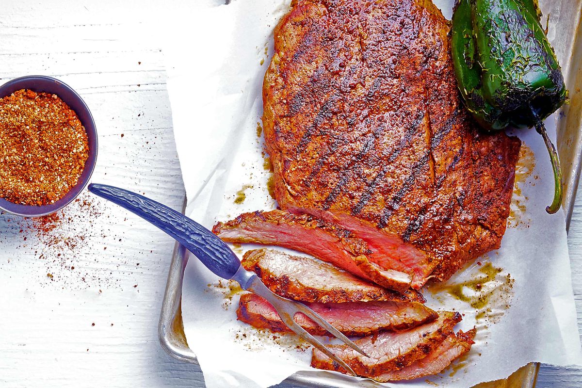 Overhead chili-rubbed flank steak sliced against the grain with a bowl of spice rub and charred jalapeño on parchment