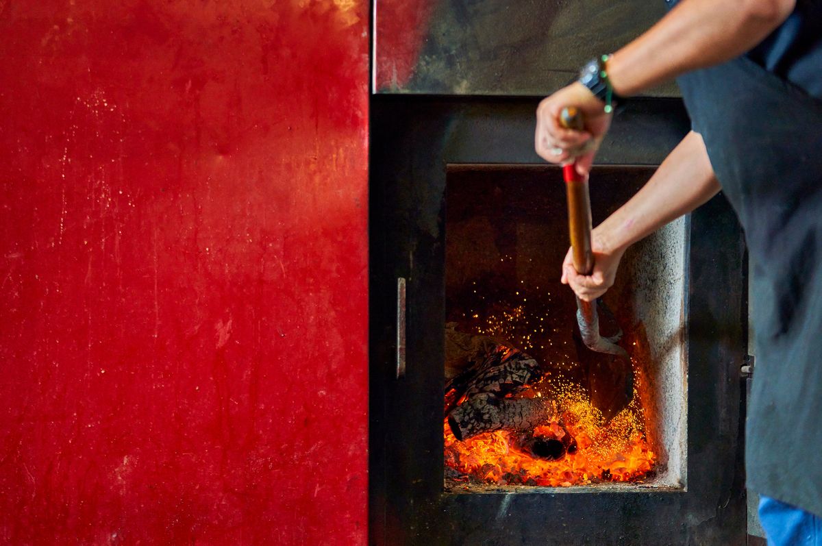 Ten50 BBQ cook shoveling glowing coals into the firebox of a wood-burning smoker against a deep red wall in Richardson, Texas