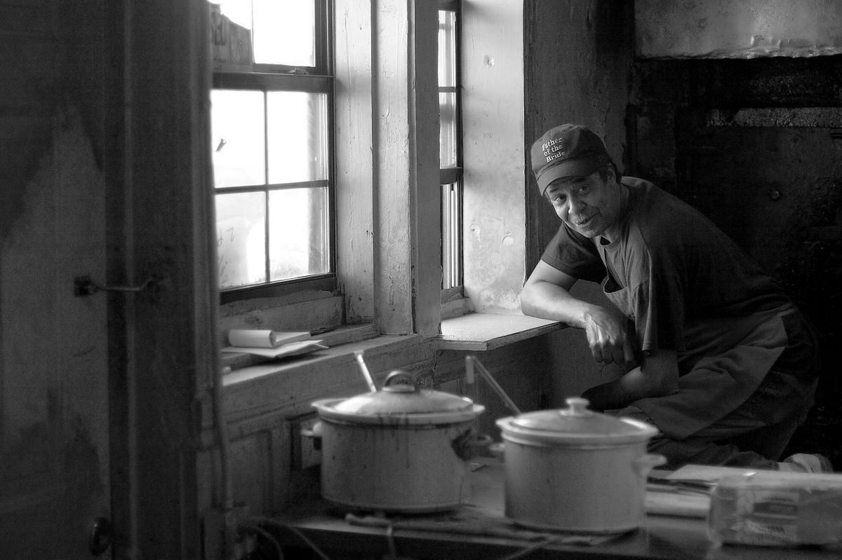 Pitmaster pausing at the window inside the Meshack's kitchen, enamel crock pots warming on the counter in black and white
