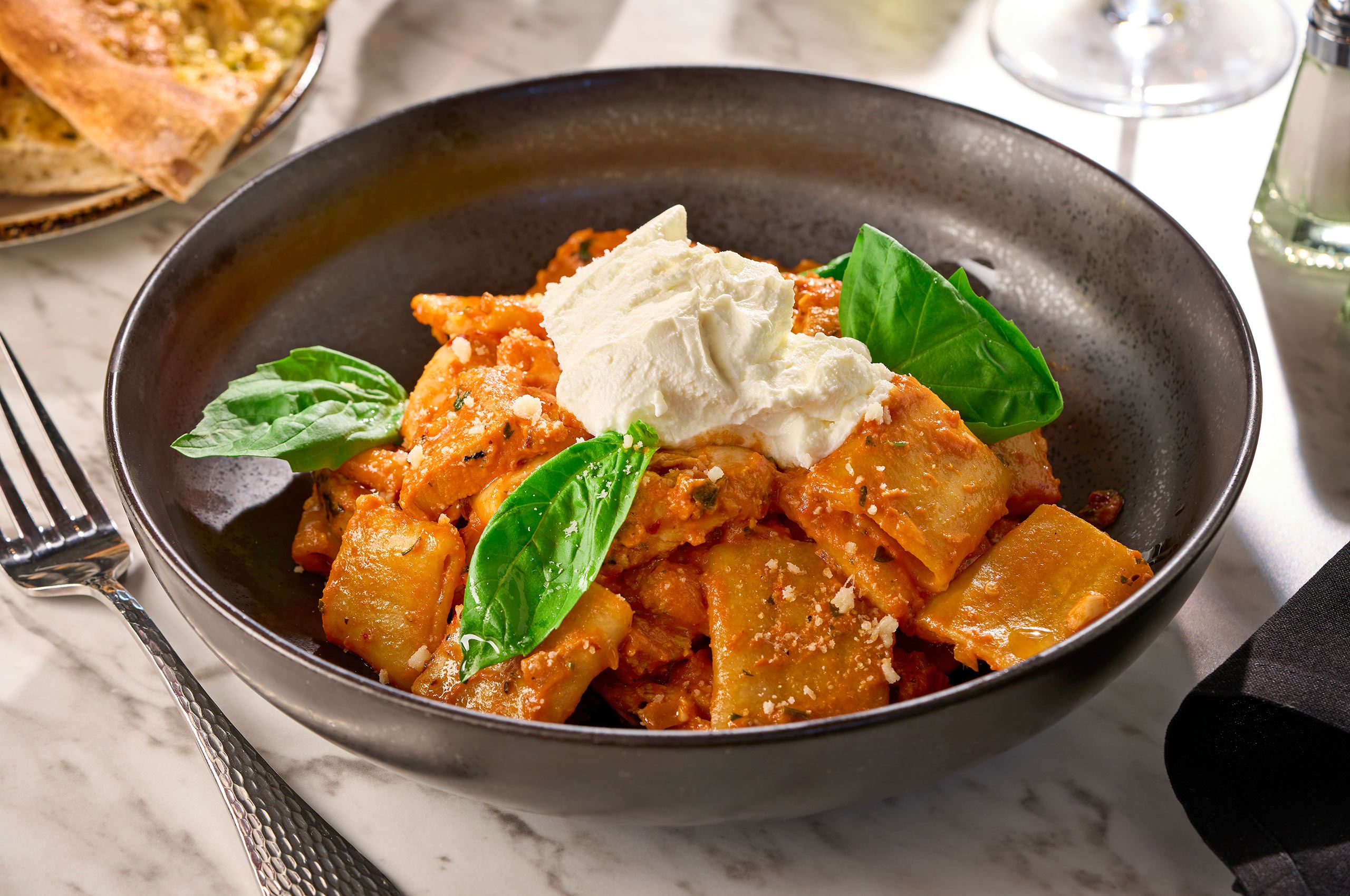 Wide ribbon pasta in a creamy tomato sauce topped with whipped ricotta and fresh basil, served in a dark bowl at Sauce & Vine
