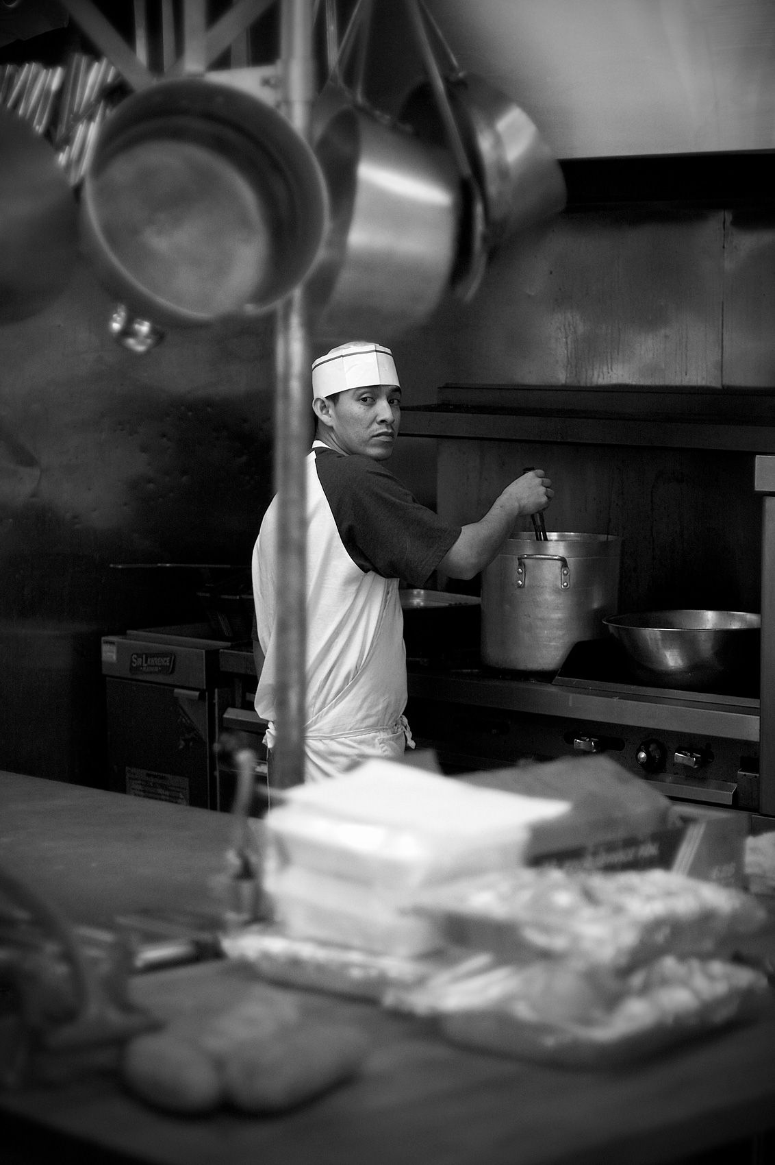 Cook glancing back from the range at Smokey's, stock pot on the burner beneath a rack of hanging pans in black and white