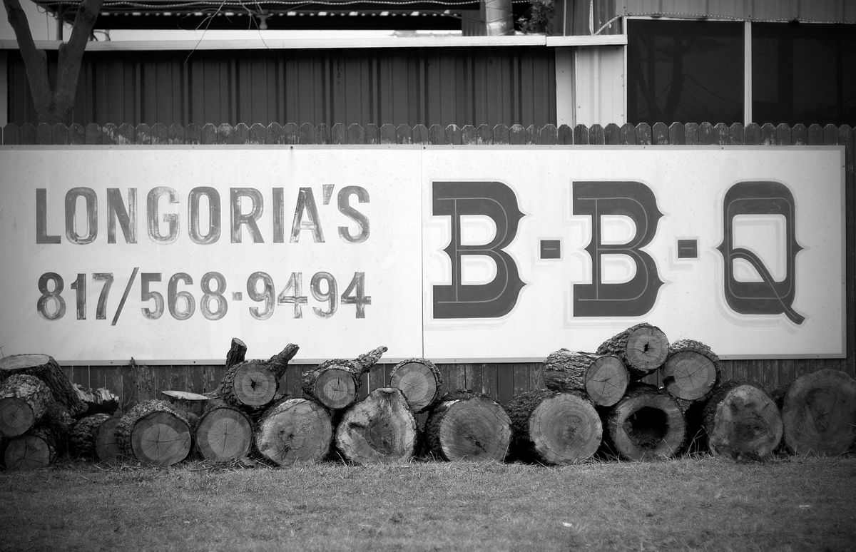Hand-lettered Longoria's B-B-Q sign above a row of cut oak logs stacked along the fence, shot in black and white
