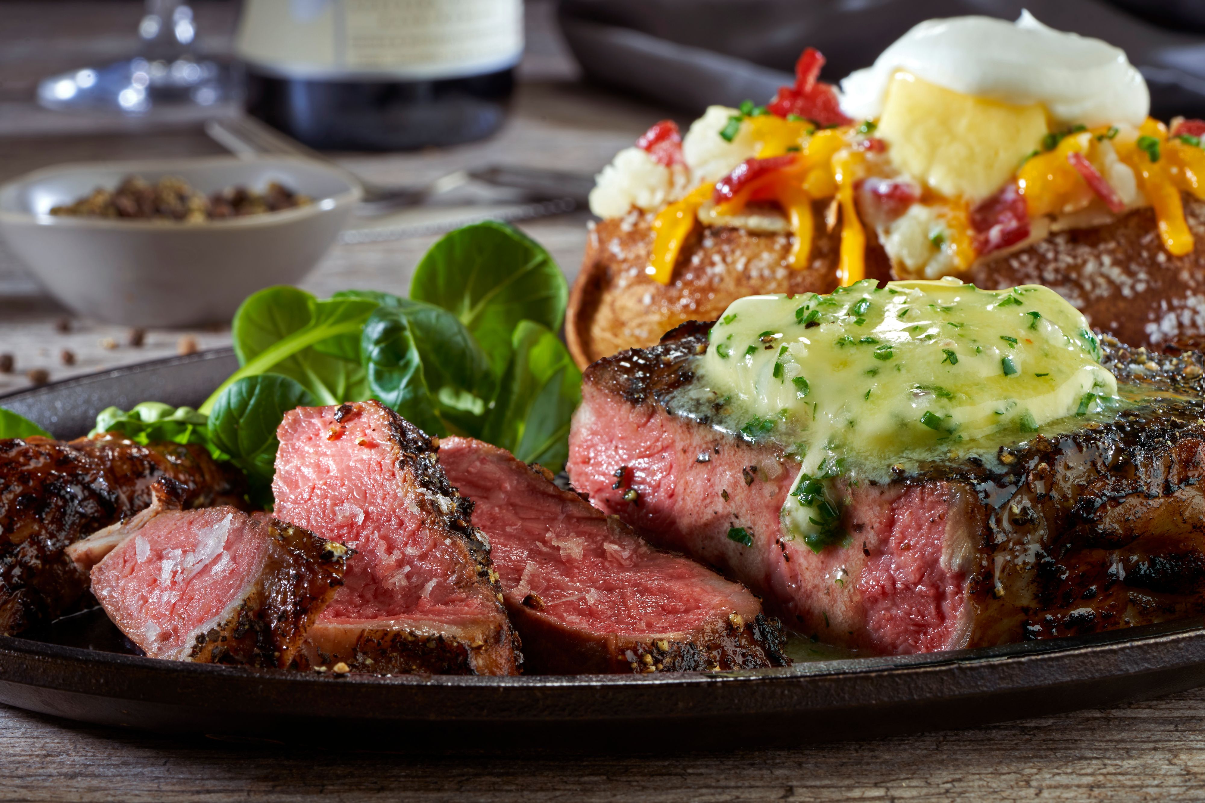 Sliced medium-rare strip steak topped with melting herb butter beside a loaded baked potato on a cast iron skillet platter