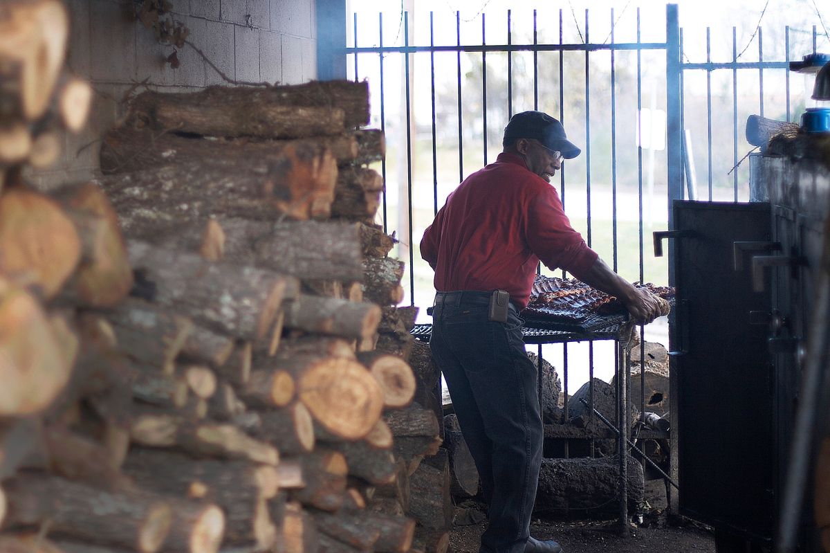 Pitmaster loading racks of ribs into the smoker at Off the Bone Barbeque, stacked firewood filling the foreground in Dallas