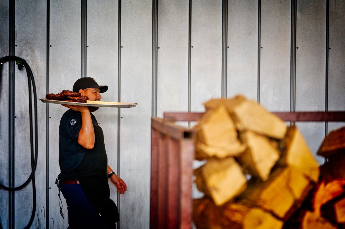 Ten50 BBQ cook carrying a tray of smoked meat on his shoulder past a stack of split wood in Richardson, Texas