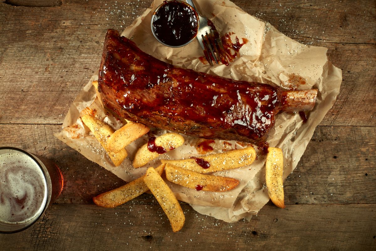 Overhead glazed barbecue rib rack with steak fries, dipping sauce, and a cold beer on butcher paper over rustic wood