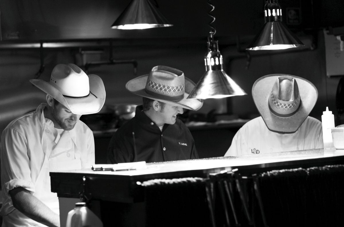 Chef Tim Love working the pass with two cooks in cowboy hats under heat lamps at Lonesome Dove in the Fort Worth Stockyards