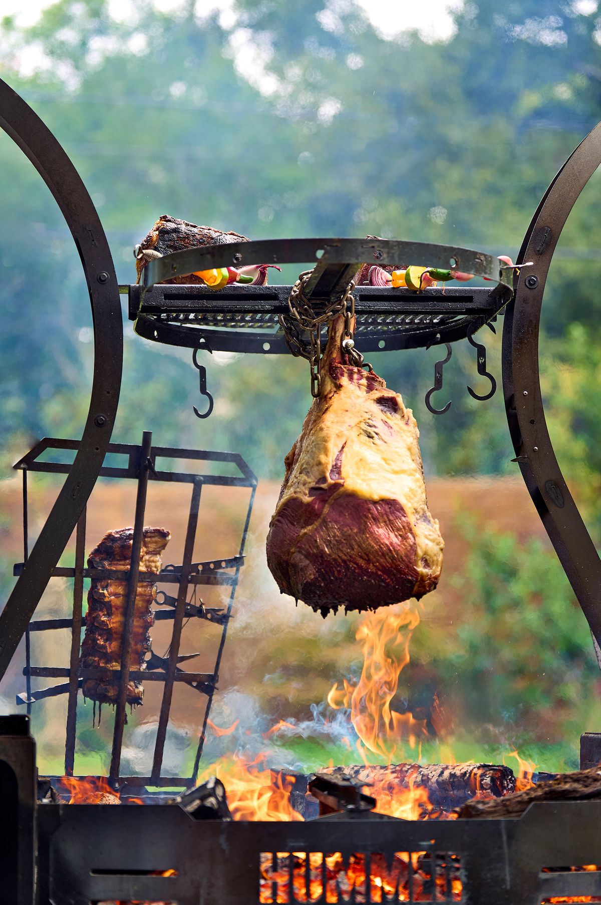 Rosewood Wagyu Beef whole cut suspended on chains over an open wood fire, vegetable skewers and ribs on the rack above