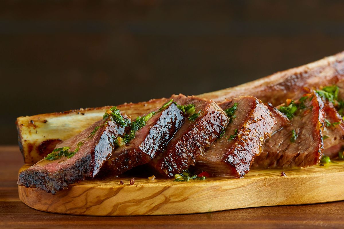 Fogo de Chão long-bone beef rib carved into thick slices and finished with chimichurri on an olive wood board