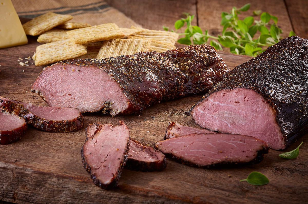 Two Ranch House mesquite smoked peppered beef roasts sliced on a rustic board with crackers, cheese, and fresh herbs