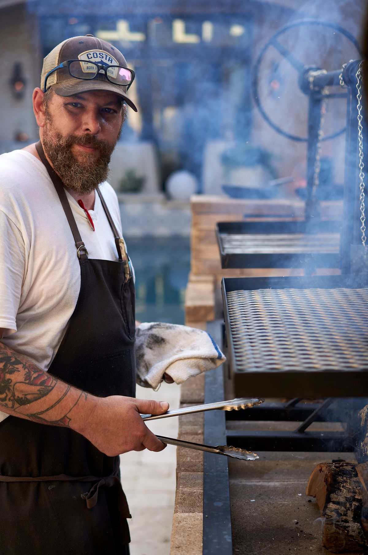Chef Brent Hammer at the open fire grill with tongs and towel in hand, smoke drifting across the cooking surface
