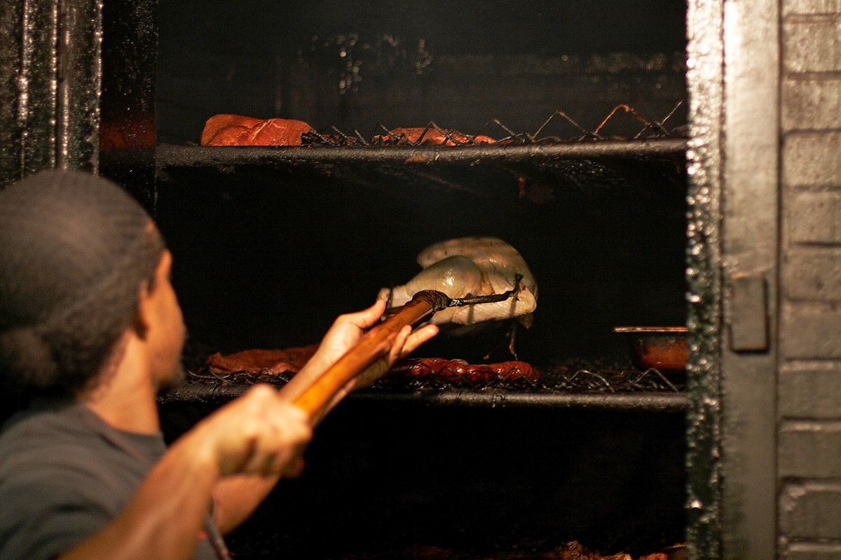 Pitmaster setting a whole chicken onto the rack inside the open brick pit at CT's, ribs and sausage links smoking below