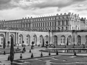 Black‑and‑white architectural study of geometric boxwoods and yews in the gardens of Château de Versailles, France, with the palace and Orangery, photographed by William Curtis Rolf. Black‑and‑white photograph of geometric boxwoods and yews in the gardens of Château de Versailles, France, with the palace and Orangery in the background under dramatic clouds.