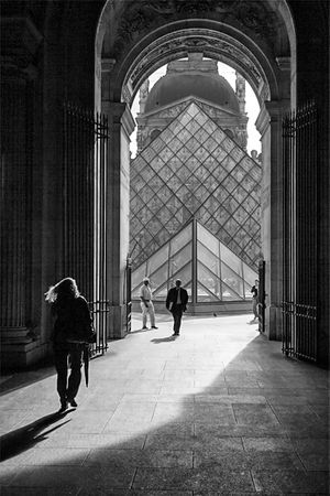 Black‑and‑white photograph of people walking through a Louvre stone arch toward the courtyard with the Glass Pyramid and surrounding classical architecture in Paris.