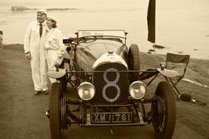 Fine-art sepia tone photograph by William Curtis Rolf capturing a 1922 Bentley 3 Litre Le Mans racer with its owners in period driving suits against a blurred Pebble Beach ocean coastline. Pebble Beach 1922 Bentley Le Mans XM 6761 Sepia Print