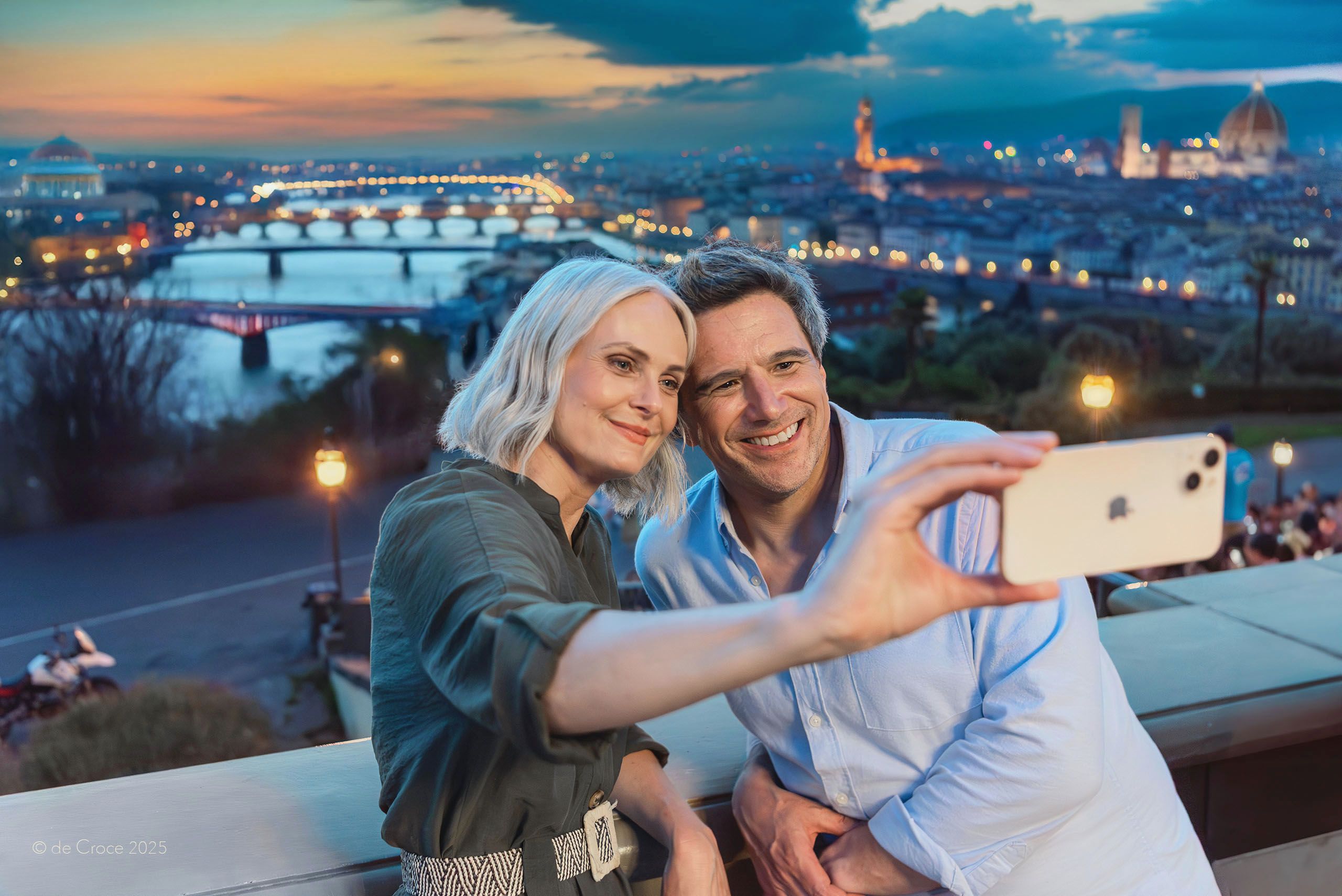 Advertising photographer captures image of professional models for travel commercial in Europe. In this image, the middle-age couple is depicted at a scenic city overlook at dusk making a selfie on their phone. Lifestyle & Travel Photography Europe