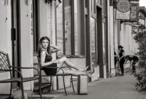 A young woman sits on bench with suitcase as restaurant workers sweep in black and white model portfolio photography. Woman on Street with Suitcase