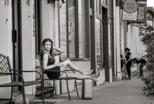 A young woman sits on bench with suitcase as restaurant workers sweep in black and white model portfolio photography. Woman on Street with Suitcase
