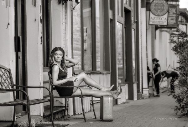 A young woman sits on bench with suitcase as restaurant workers sweep in black and white model portfolio photography. Woman on Street with Suitcase