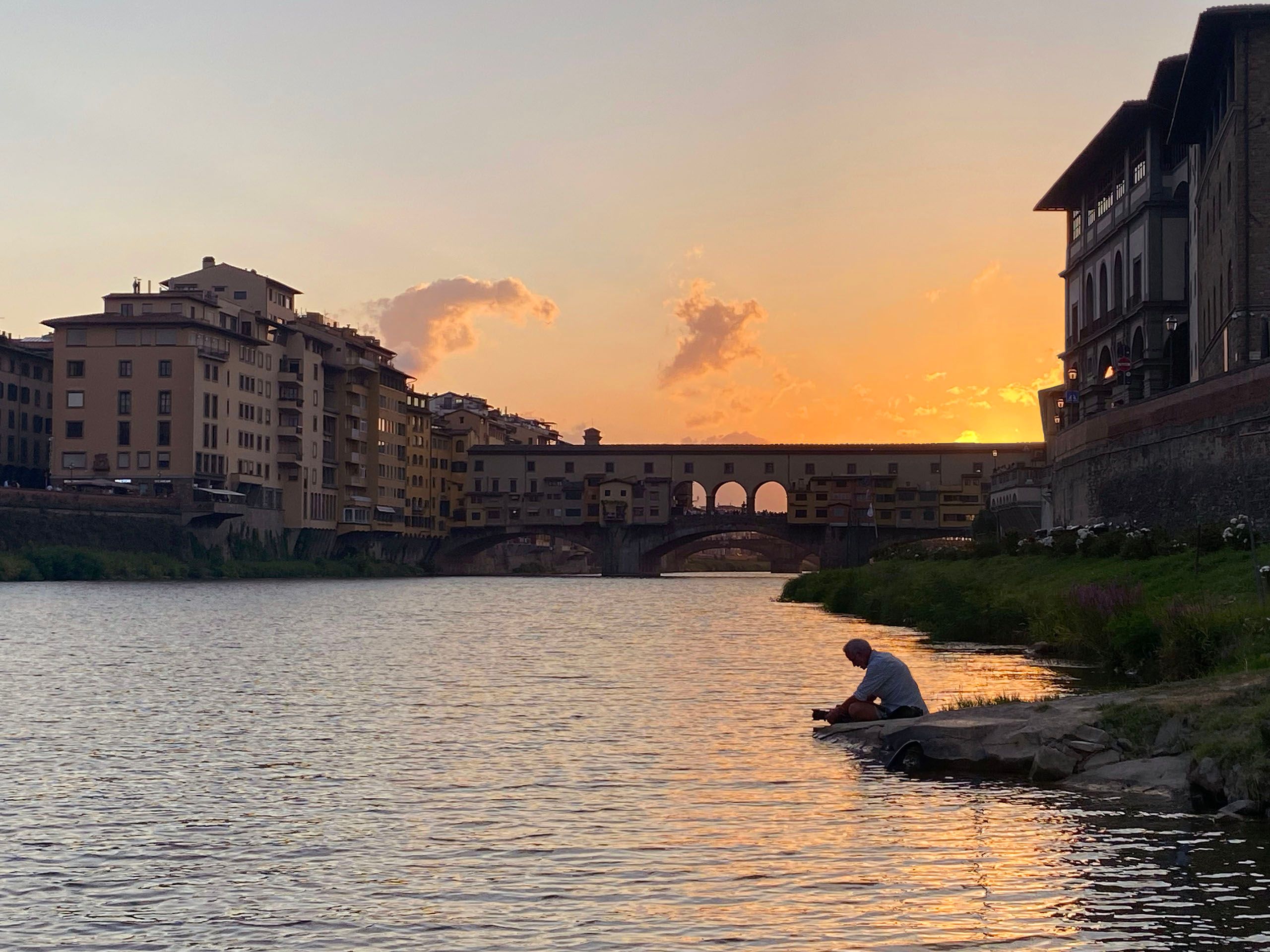 Travel photographer, Edward DeCroce, at work with camera on the bank of The Arno River in front of Ponte Vecchio, Florence Italy. The week long Italian endeavor was the making of a video commercial photoshoot for Globus Adventures, based in Switzerland. The aim of the 30 second advertisement was to contrast two styles of travel. One couple who booked their European sojourn through Globus Adventures had an experience of a lifetime. They took cooking classes, rode horses at a country castle, avoided long lines and, as seen in this travel portfolio, boated on the Arno River through Florence. The another couple started their trip with all the best intensions but became frustrated with logistics and found themselves worn out by the hassles of travel. DeCroce was hired as the stills photographer. This photo was made by professional sound-man Giacomo Verranda. July 2025. Travel Photographer DeCroce Ponte Vecchio