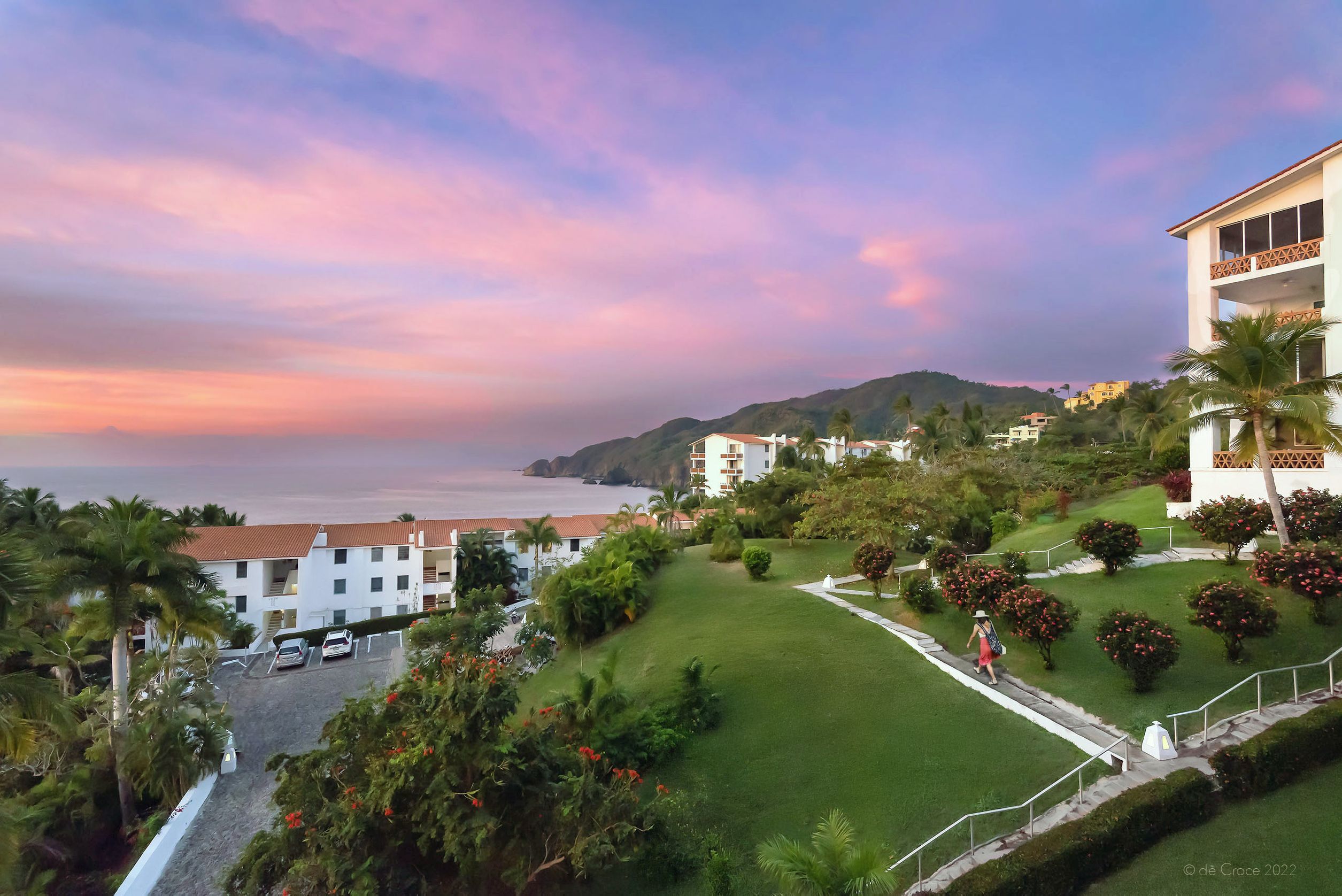Commercial photographer based in Denver Colorado traveled to Colima Mexico for vacation lifestyle photoshoot. The scene here shows a woman in beach attire walking on pathway of luxurious condo on the Pacific shore under pink and orange dawn sky. Commercial Photography - Travel Advertisement Condos Colima