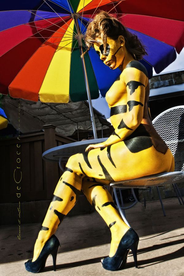 Wearing nothing but paint, this model in high heels (painted as a bee) poses outside at cafe for Denver people photographer. Body Paint Art Photography Denver
