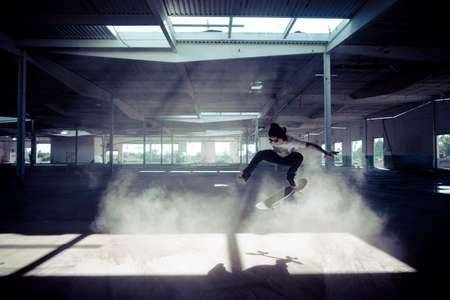 Danny Scher. Carlsbad, CA in the air on his skateboard in a moody dusty warehouse. Skateboarder ollies in dusty warehouse