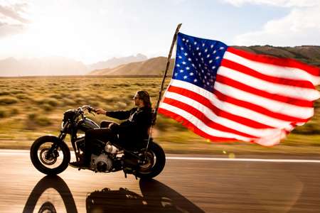 Jeremy Jones. Jackson, WY. Crusing on a Harley Davidson with a giant American flag in backlit rain and mountains. Jeremy Jones riders his motorcycle in pouring backlit rain