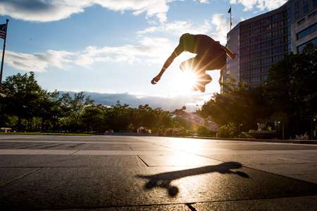 Mike Nalls. Washington, DC flipping board during sunset with large shadow of skateboard on the ground. Skateboarder flips board through the sun