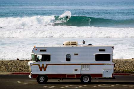 Surfer turns and sprays on a wave in California. An RV is parked in the parking lot in the foreground. No one else is around Surfer turns on a wave in front of Winnebago RV