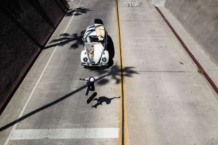Danny Scher. Oceanside, CA.. Crusing down the street doing a trick while a surf mobile follows him with surfboard on the roof and a palm tree shadow on the ground A skateboarder skates down street with a VW Bug following him