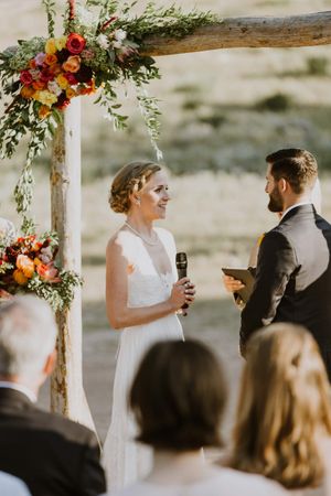 Bride and Groom Exchanging Vows in Front of Rustic Arch with Peach and Read Roses Ceremony